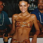 Woman in leopard print bikini holding a beer at a bar with two men behind her.