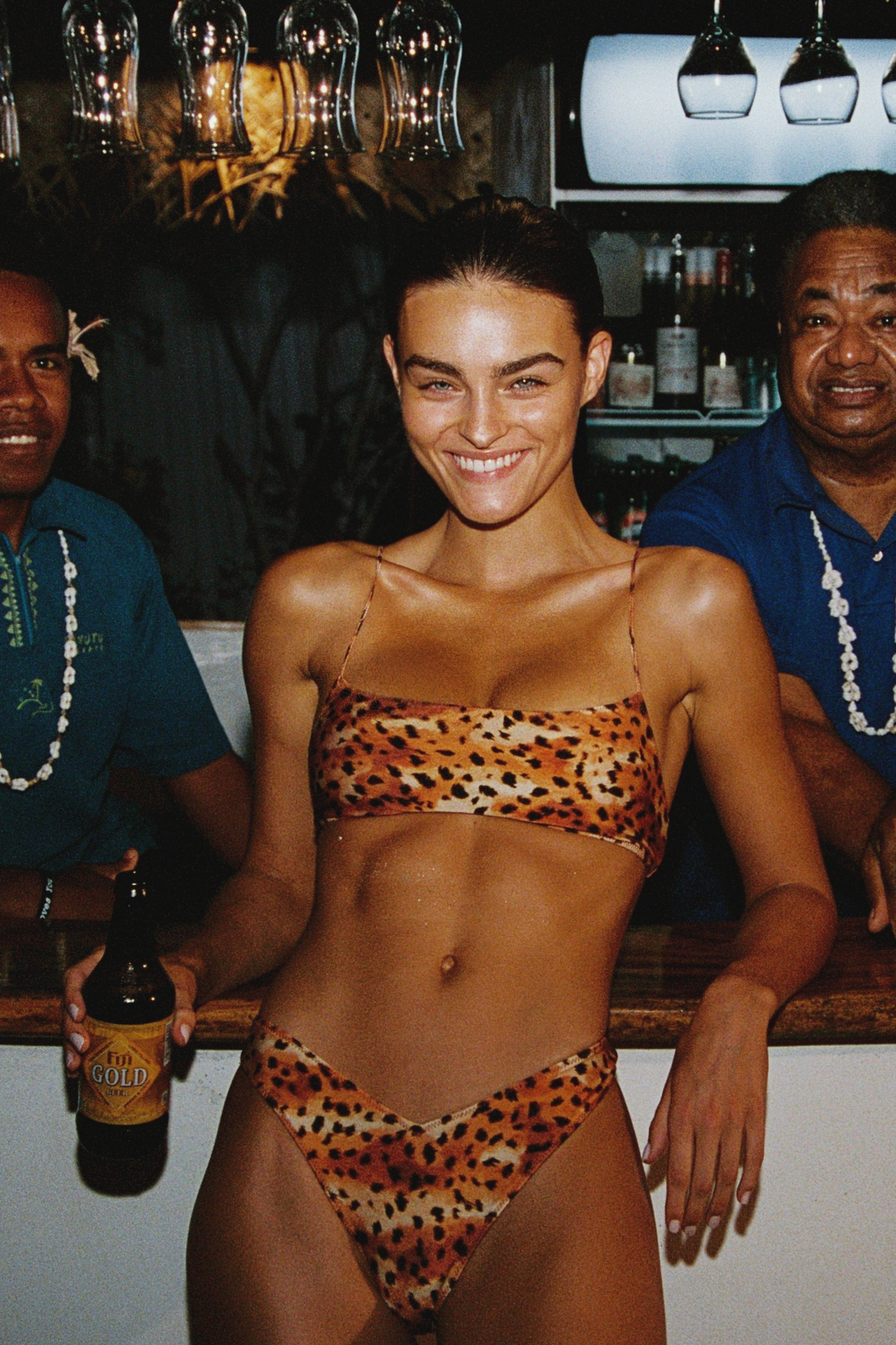 Woman in leopard print bikini holding a beer at a bar with two men behind her.