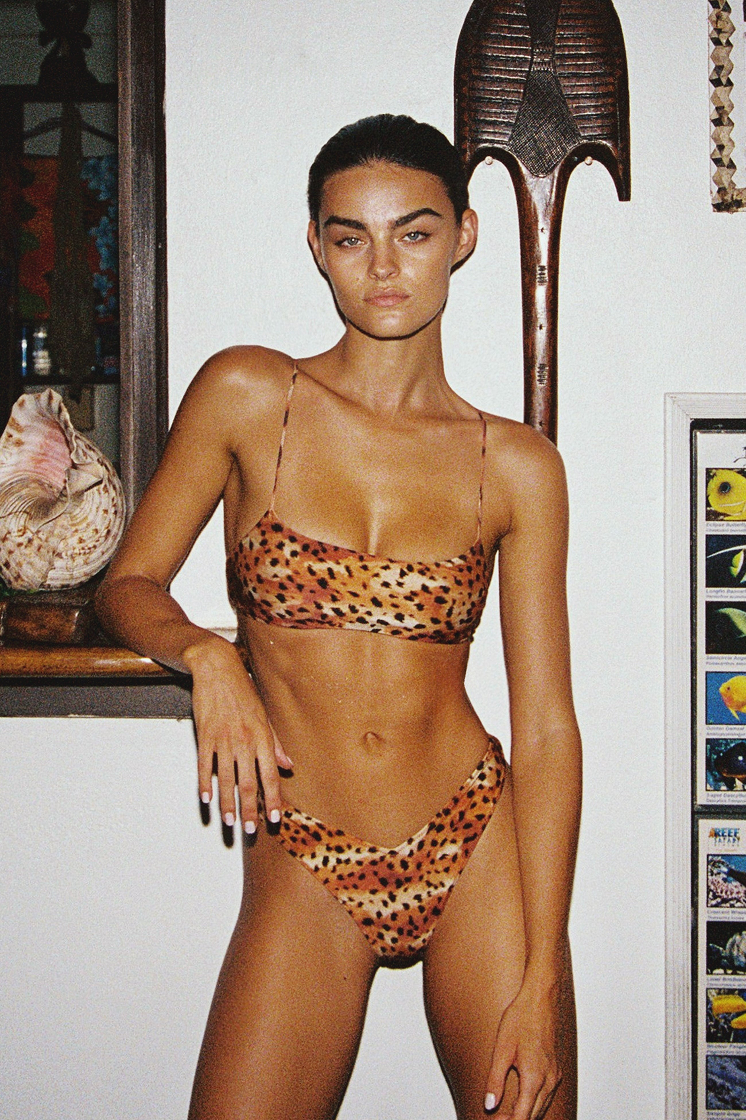 Woman wearing a leopard print bikini in an indoor setting with a shelf and decorative items.