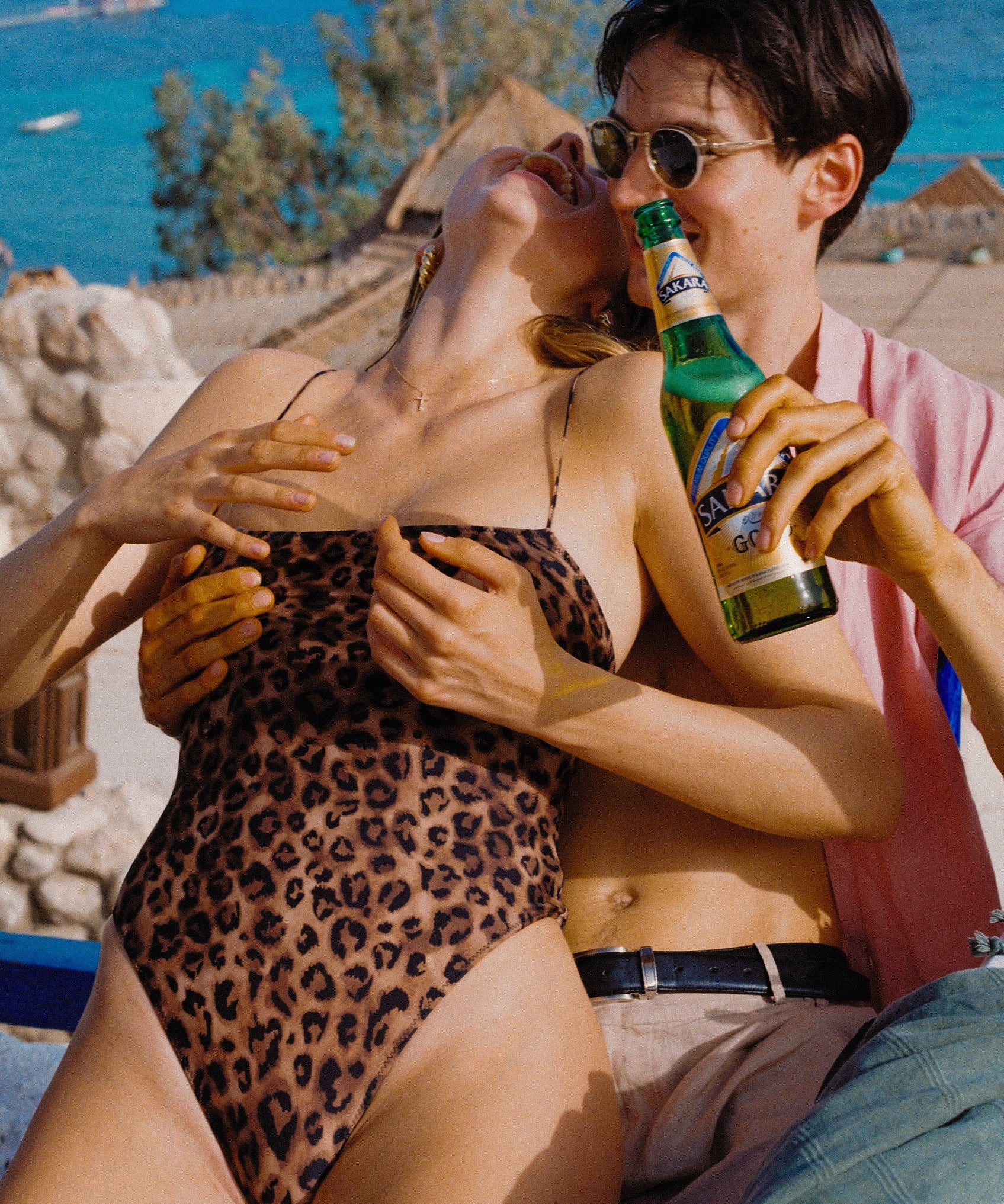 A person in a leopard print one-piece swimsuit is sitting close to another person who is holding a beverage bottle. They appear to be at a beach or poolside setting.